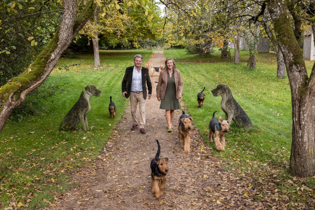 Ein Paar geht mit mehreren Hunden auf einem von Bäumen gesäumten Weg durch einen herbstlichen Park. Einige Hunde sind lebendig, während andere wie Skulpturen aussehen.
